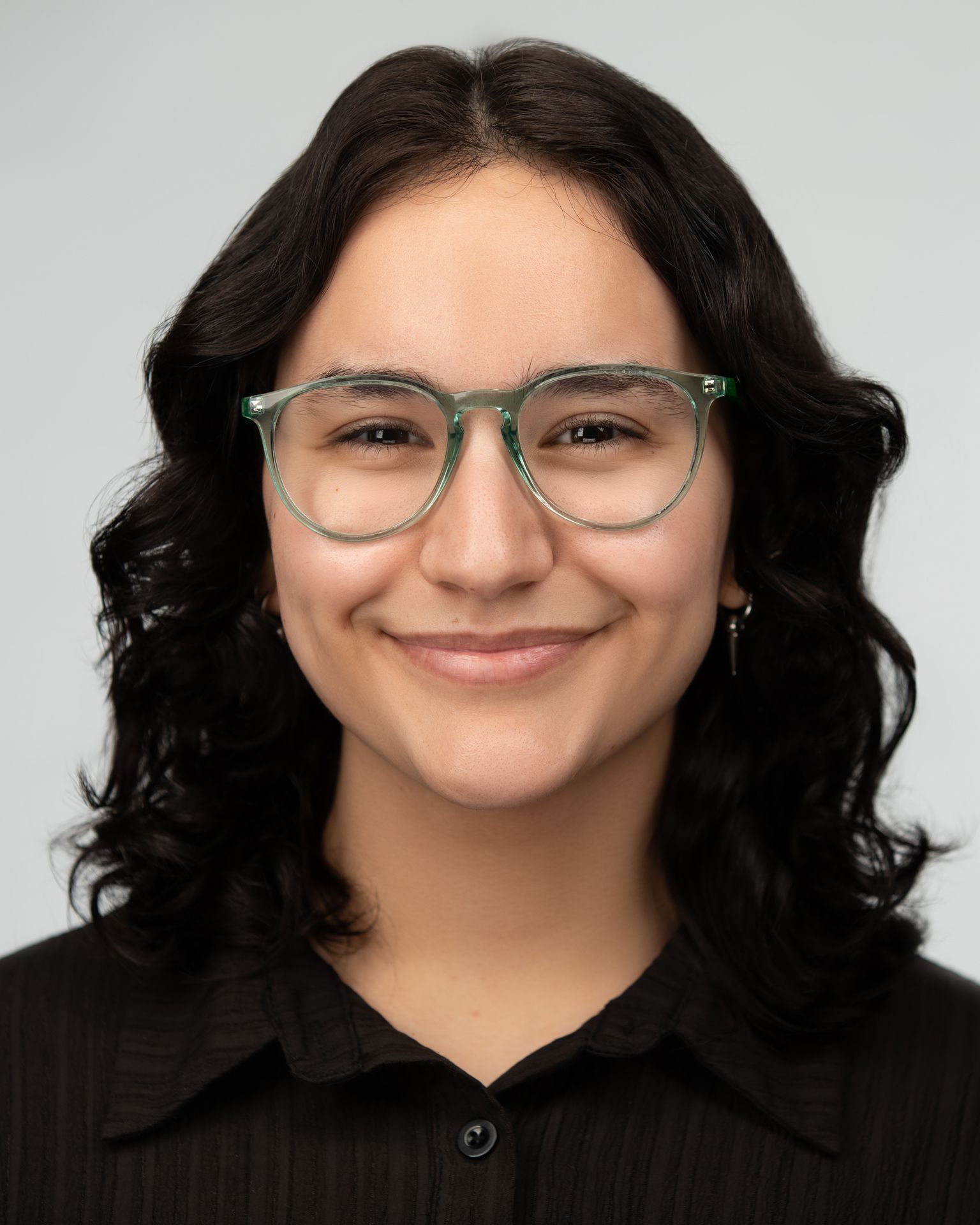 Professional headshot of Melinda Estrada wearing a dark green crew-neck top with geometric dangling earrings. The individual has short, tightly curled hair. The background is solid black, creating a professional studio portrait setting.
