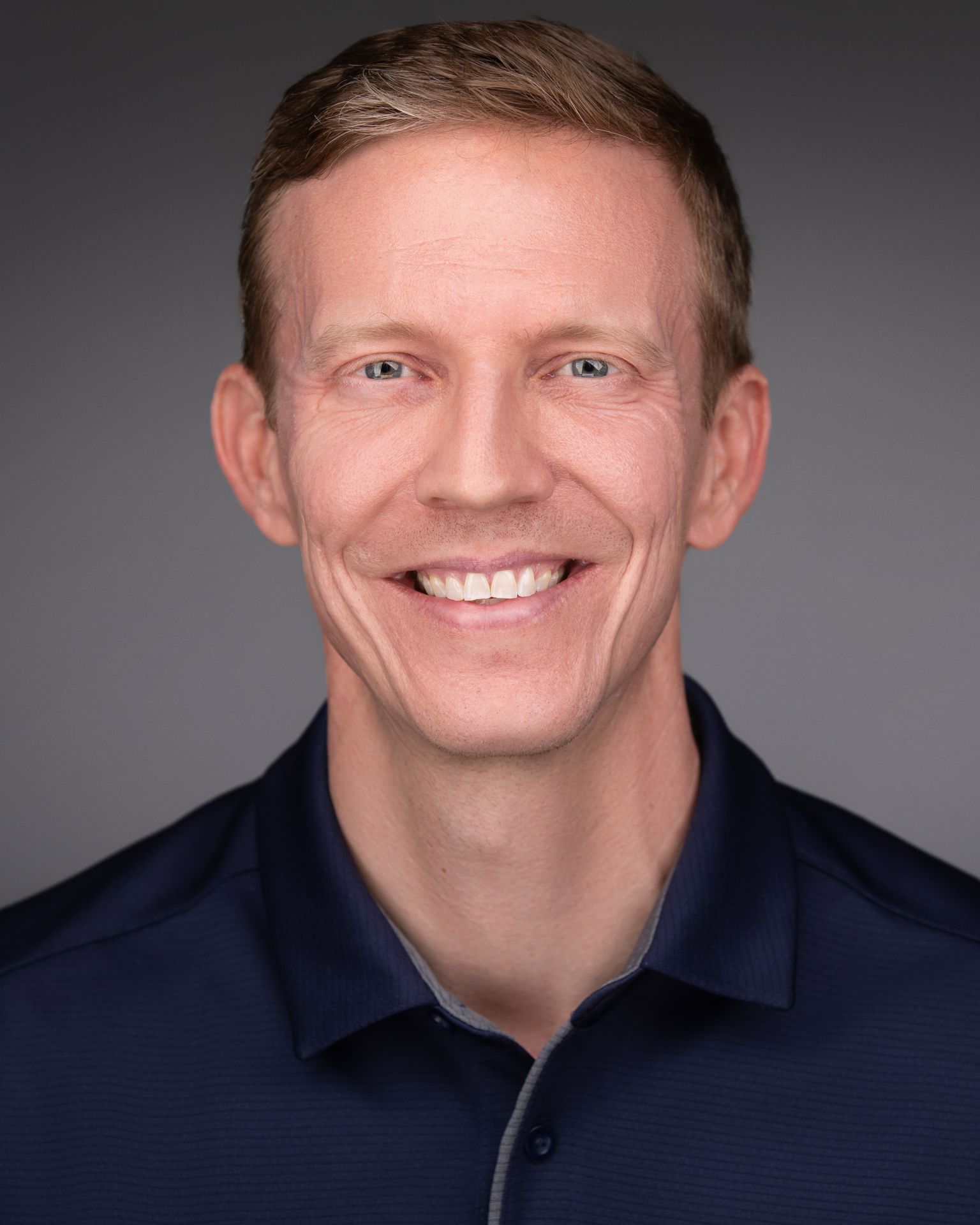 Person wearing a dark blue collared shirt with a contrasting inner lining, featuring button details. The individual has short, wavy dark hair and is wearing small hoop earrings in both ears. The background is solid black, creating a studio-style portrait look.