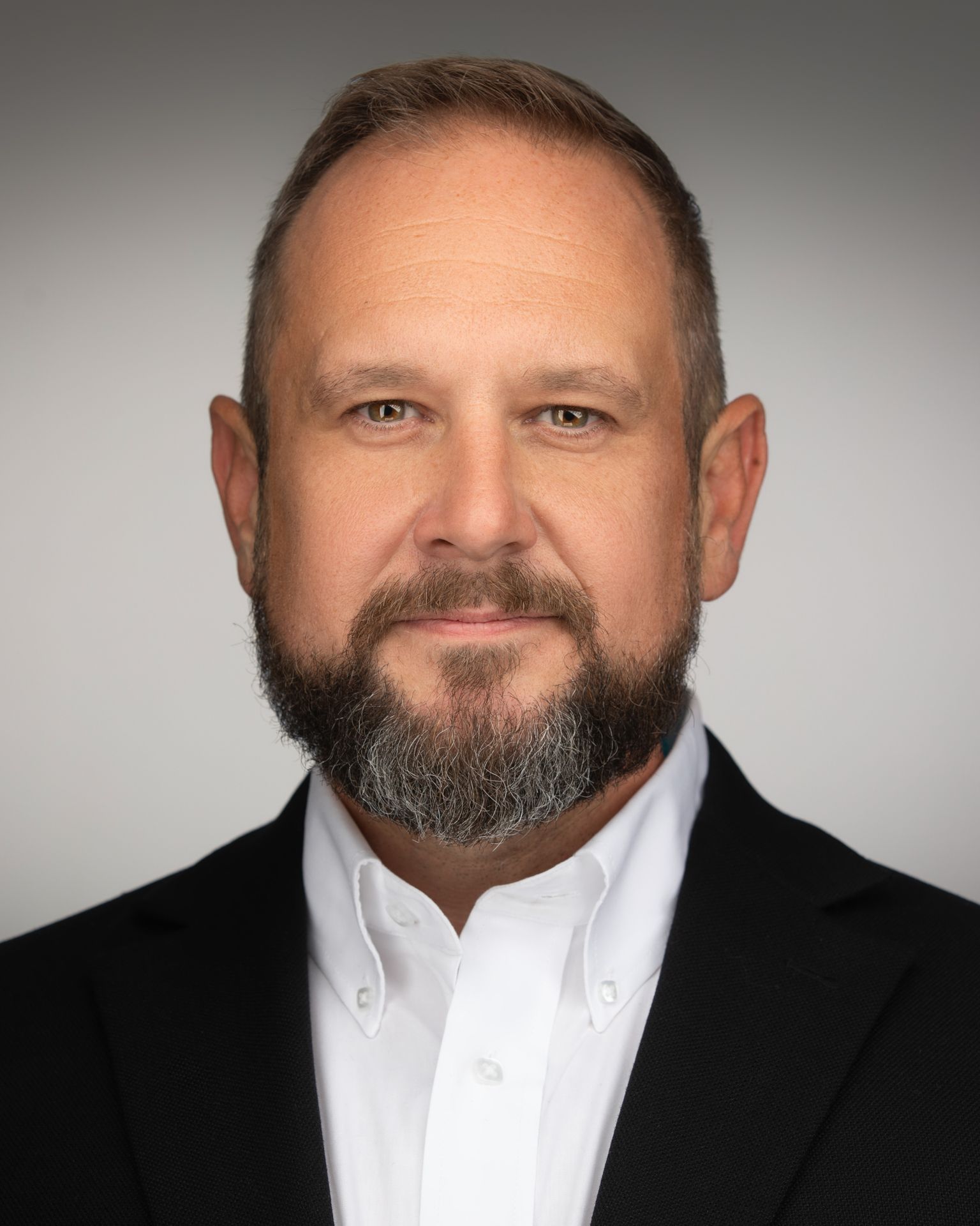 Professional Headshot of Lauren Mendez with shoulder-length, layered brown hair wearing a black blazer over a light gray top. A delicate gold necklace with a rectangular pendant is visible. The background is solid black, creating a professional studio portrait setting.