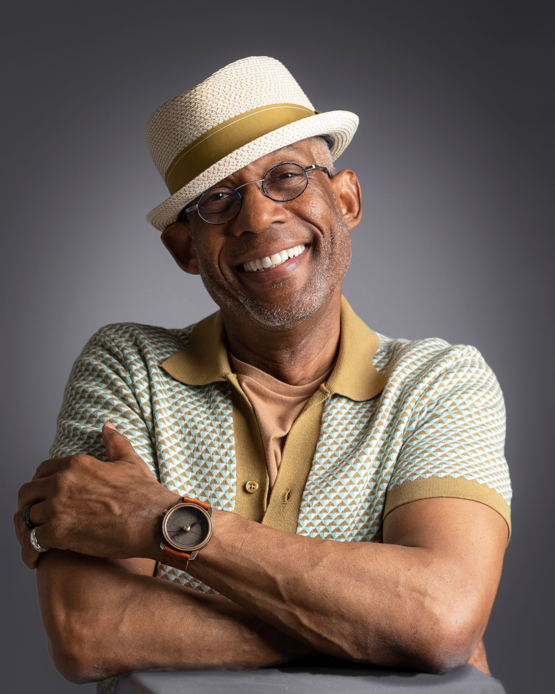 Person wearing a light-colored straw hat, short-sleeve patterned knit shirt with tan collar and cuffs, and a tan undershirt. Arms are crossed, showing a wristwatch with a dark face and orange accents, along with rings on both hands. Neutral gray background.
