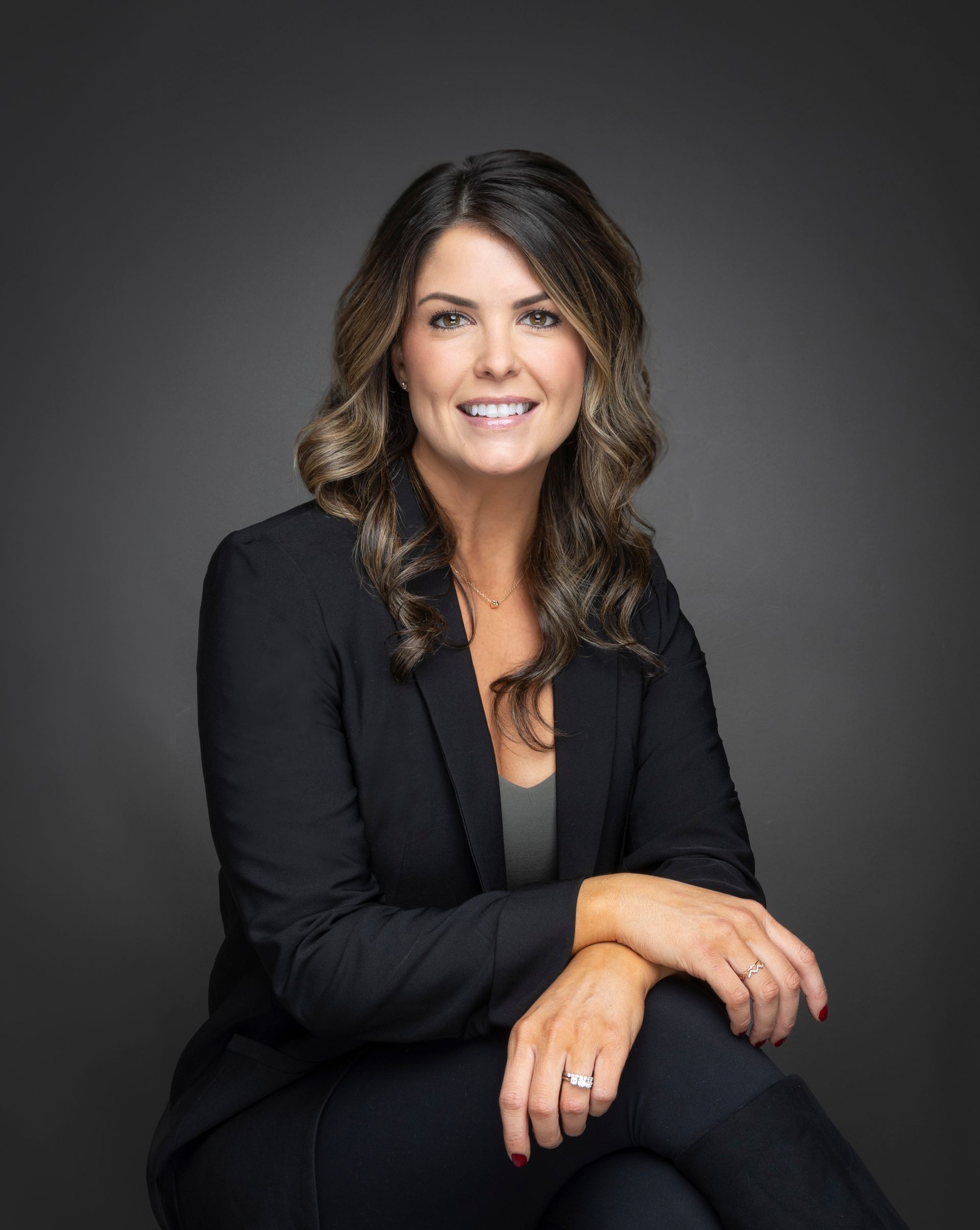 Person seated against a dark gray background, wearing a black blazer over a dark top, with wavy shoulder-length hair. Hands are resting on the lap, showing rings on both hands and neatly manicured nails.