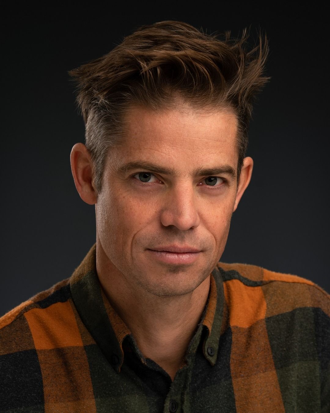 Man with spiky brown hair, wearing a green and orange plaid shirt, looking directly at the viewer.