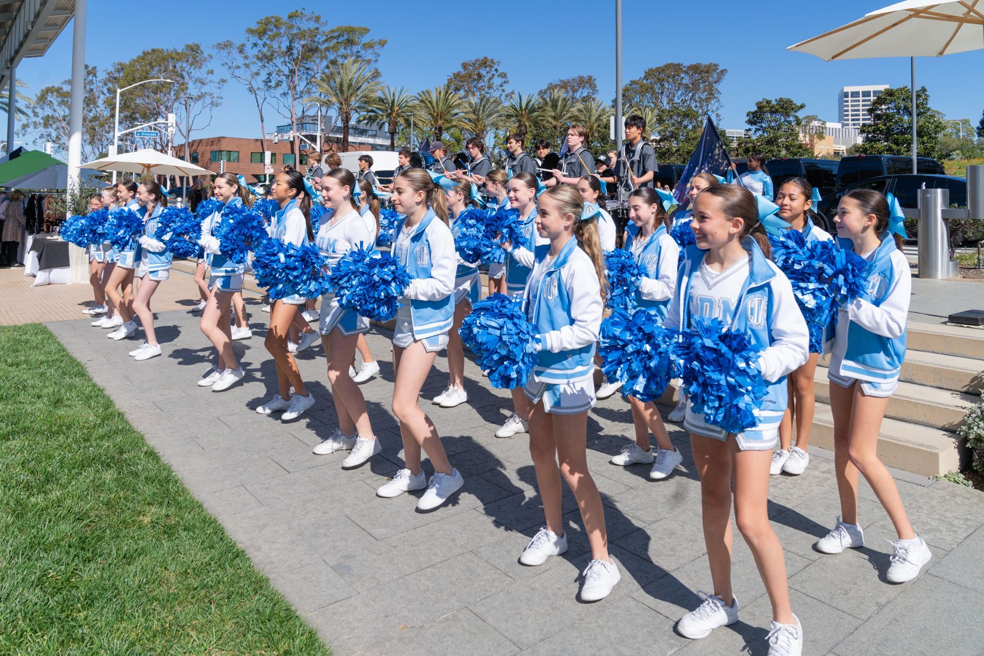 Cheerleaders in blue and white uniforms performing with pom-poms at an outdoor school spirit event surrounded by spectators.