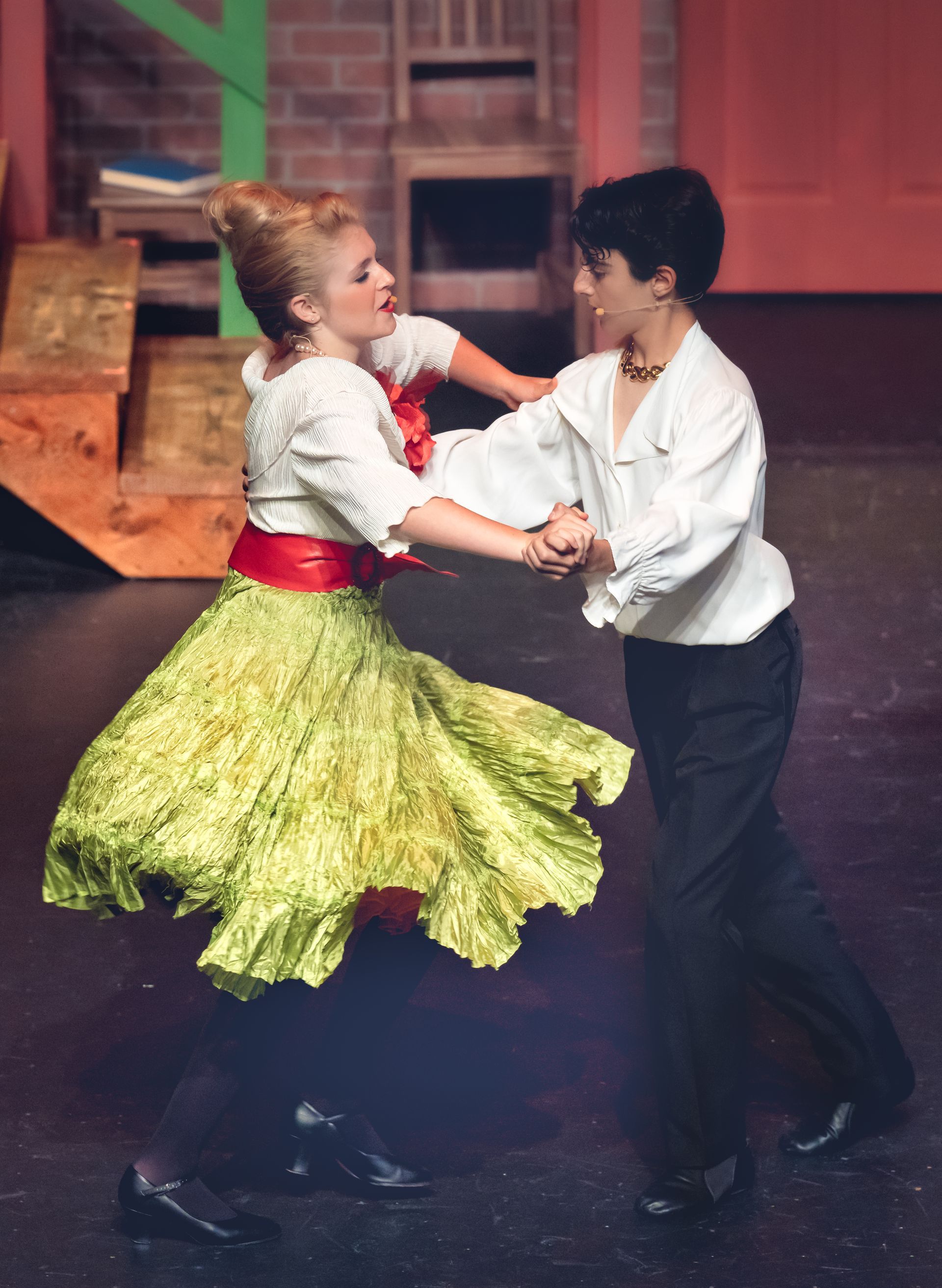Two dancers in vibrant period costumes performing on stage with wooden props and a colorful brick backdrop.