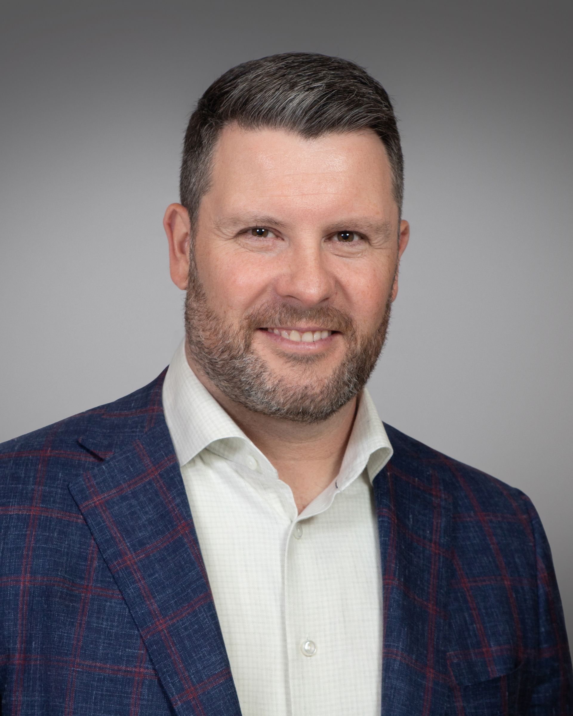 Corporate headshot featuring a person wearing a blue plaid blazer over a light-colored dress shirt against a neutral gray background.