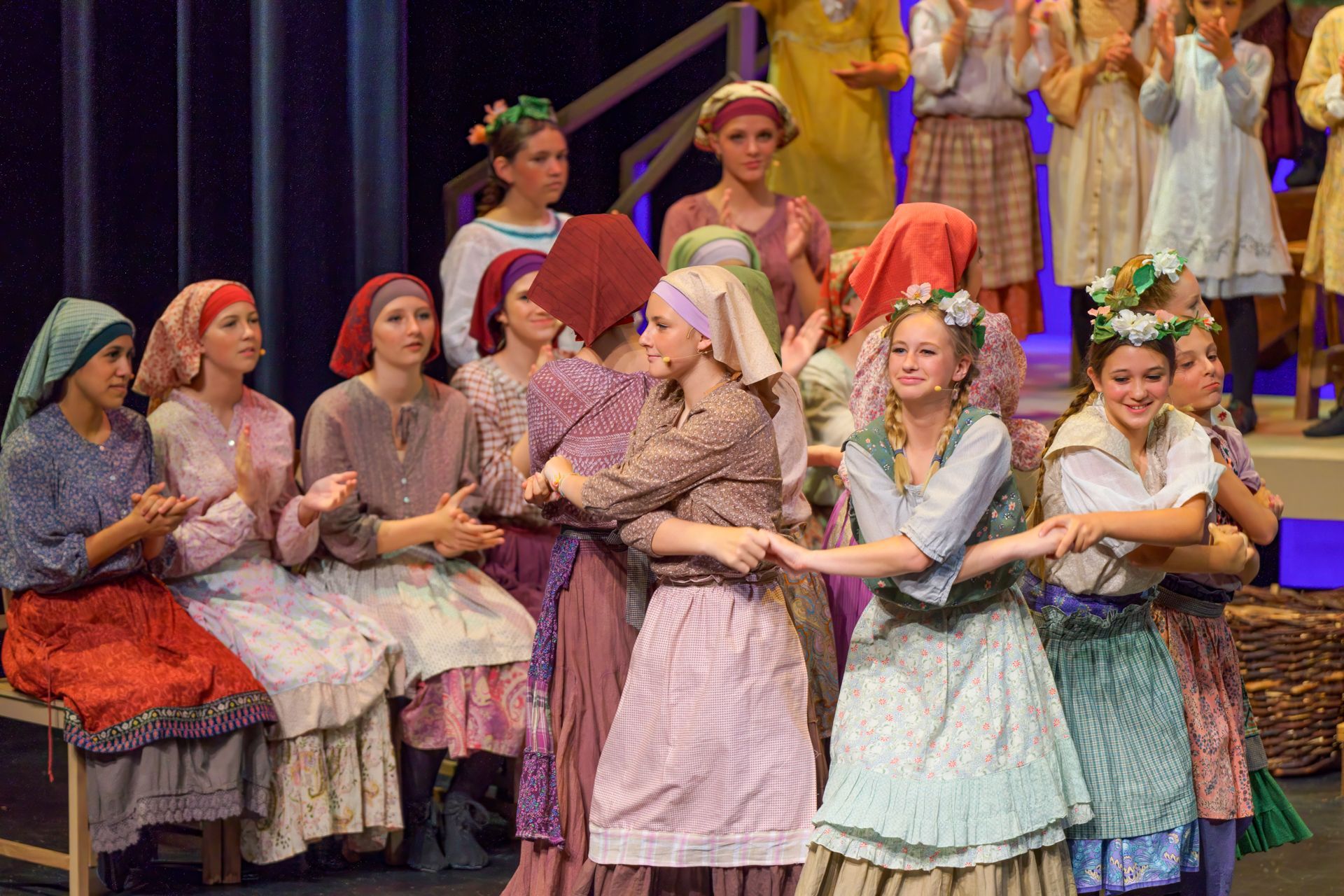 Actors in traditional folk costumes—long skirts, aprons, headscarves, and floral crowns—perform a lively dance with clapping on stage.
