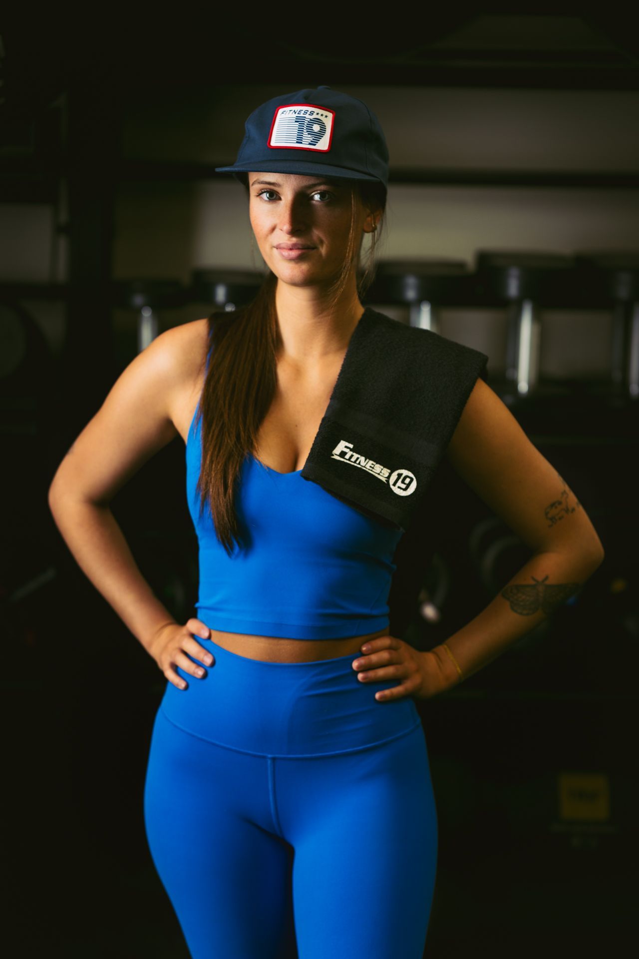 Two people standing outdoors on a balcony overlooking water and houses. Both are wearing athletic clothing with weighted shoulder straps labeled “Fitness 19.” One person is dressed in a pink sports outfit and a dark cap, while the other is in a black sleeveless top, gray jogger pants, and a backward cap. Trees and waterfront homes are visible in the background under bright daylight.