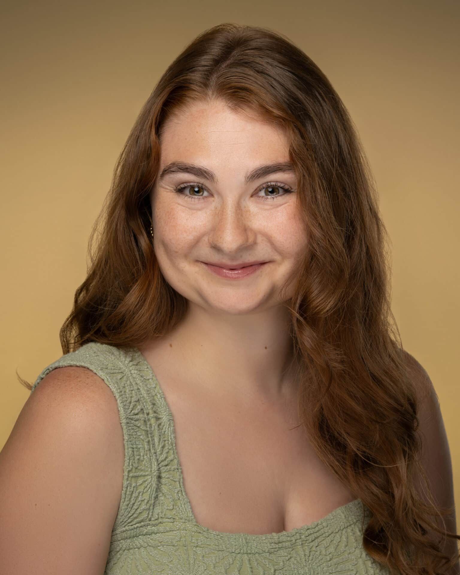 Woman with long auburn hair smiles, wearing a green textured top, against a golden backdrop.