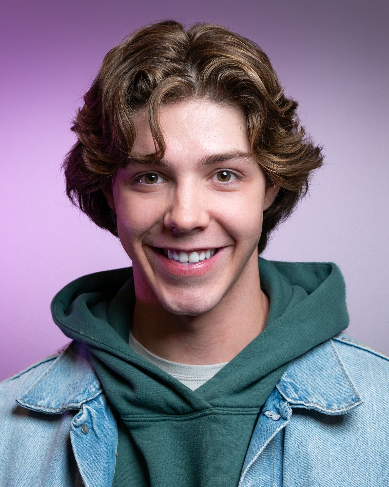 Young person smiling, wearing a denim jacket over a green hoodie, against a purple backdrop.