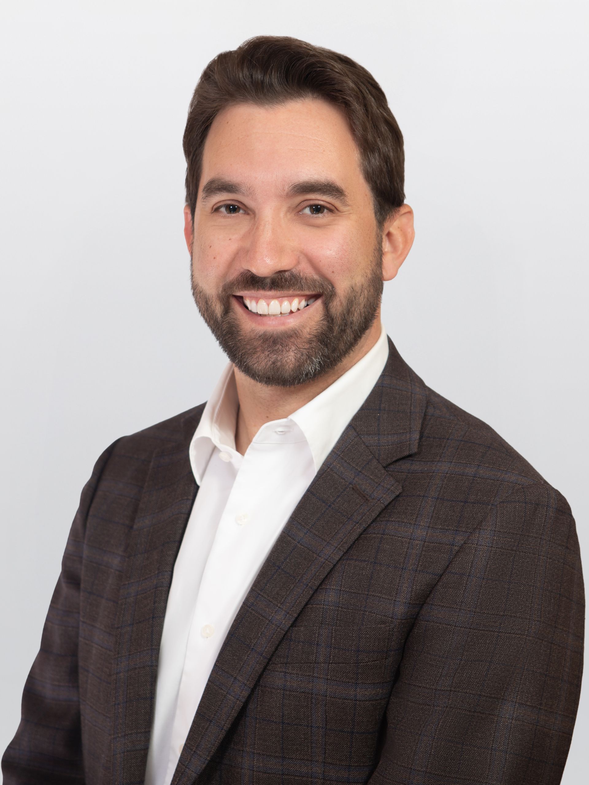 Corporate headshot featuring a person in a dark plaid blazer and crisp white dress shirt against a light gray background.