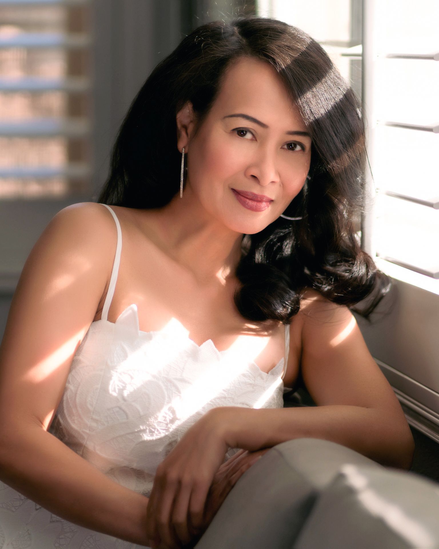 Elegant woman in a white dress by a sunlit window, showcasing natural beauty and serene expression. Art of Portraits branding included.