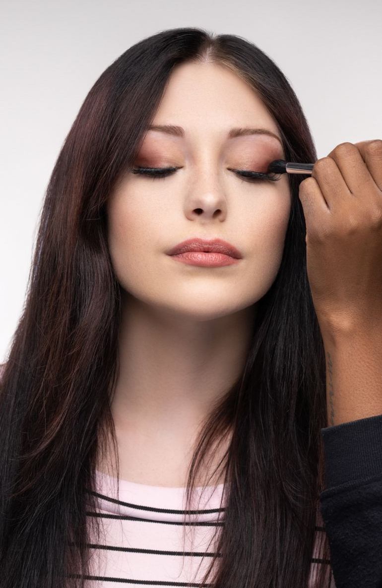 Behind-the-scenes shot of a makeup artist applying cosmetics with a brush to a person wearing a light pink top with black stripes. The scene is set against a plain white studio background, highlighting the preparation process for a professional photo shoot.