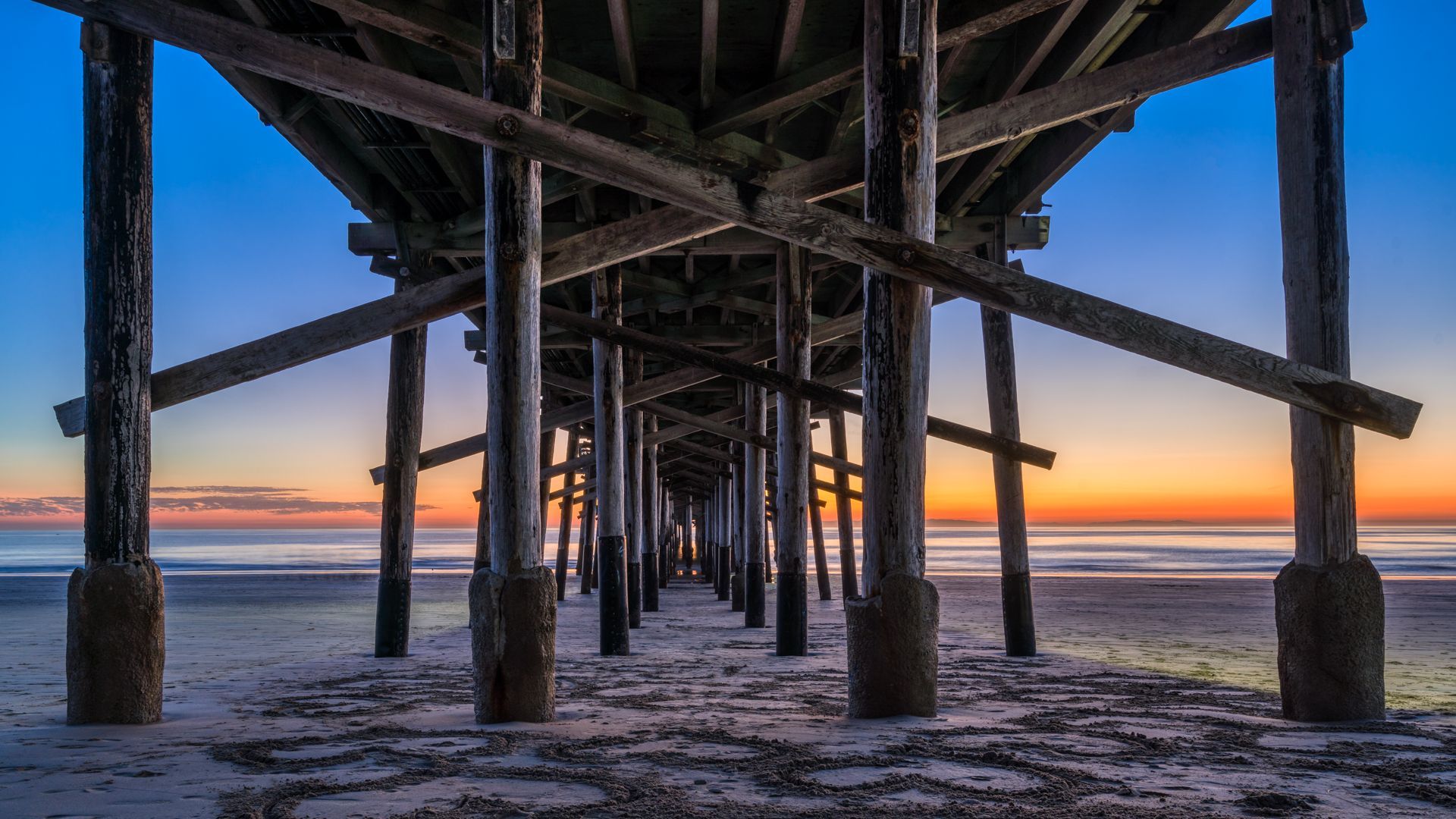 View from underneath a Newport Beach pier at the beach during sunset, with symmetrical beams and pillars leading toward the horizon over calm ocean waves.