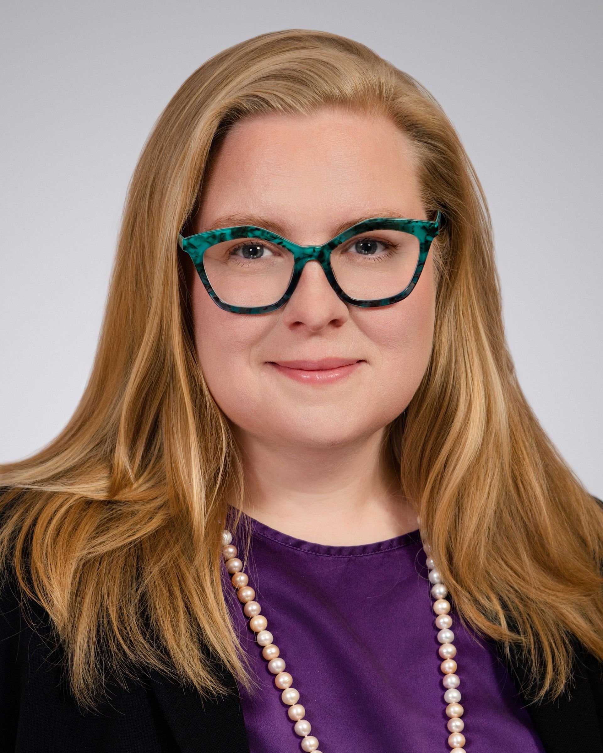 Professional portrait featuring straight, shoulder-length blonde hair, purple top, black blazer, and a pearl necklace.