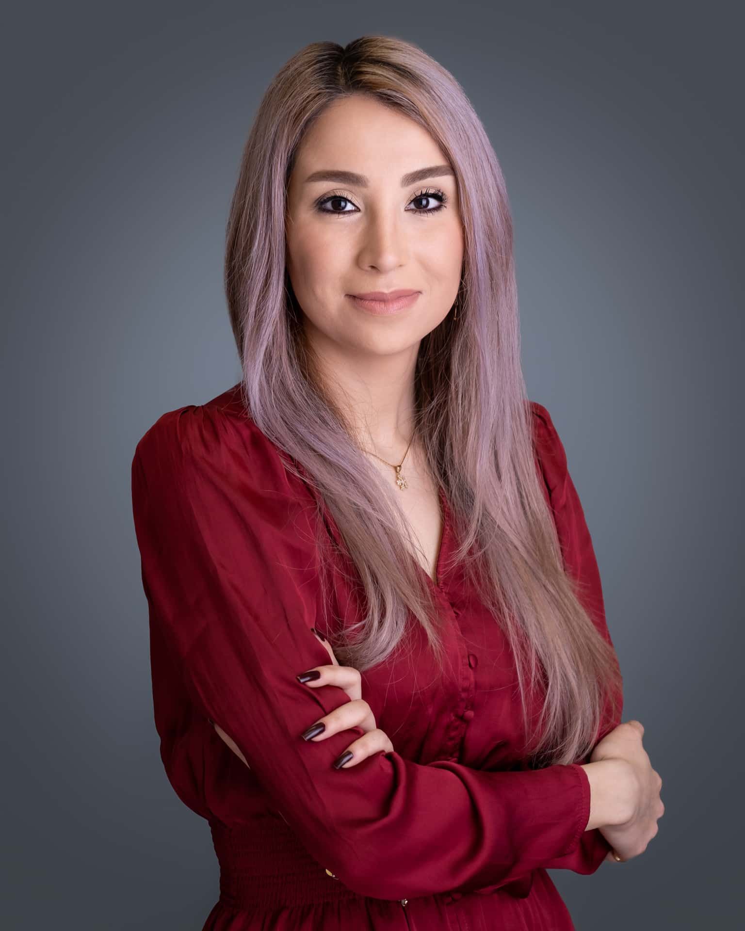 Woman with pinkish hair wearing a red dress, arms crossed, looking at the viewer. Grey background.