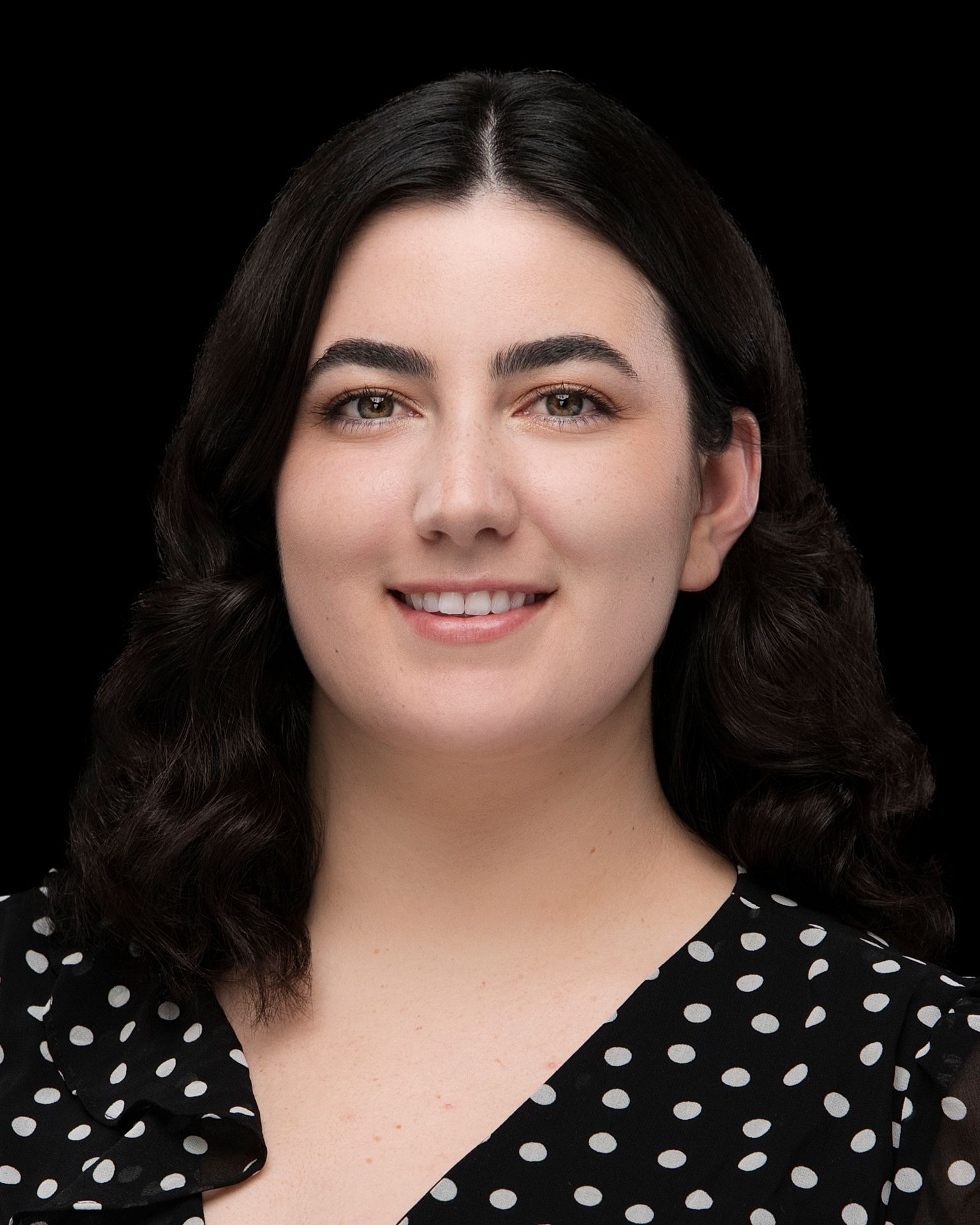 Professional headshot of a team member wearing a black blouse with white polka dots, featuring wavy dark hair parted in the center, set against a solid black background.