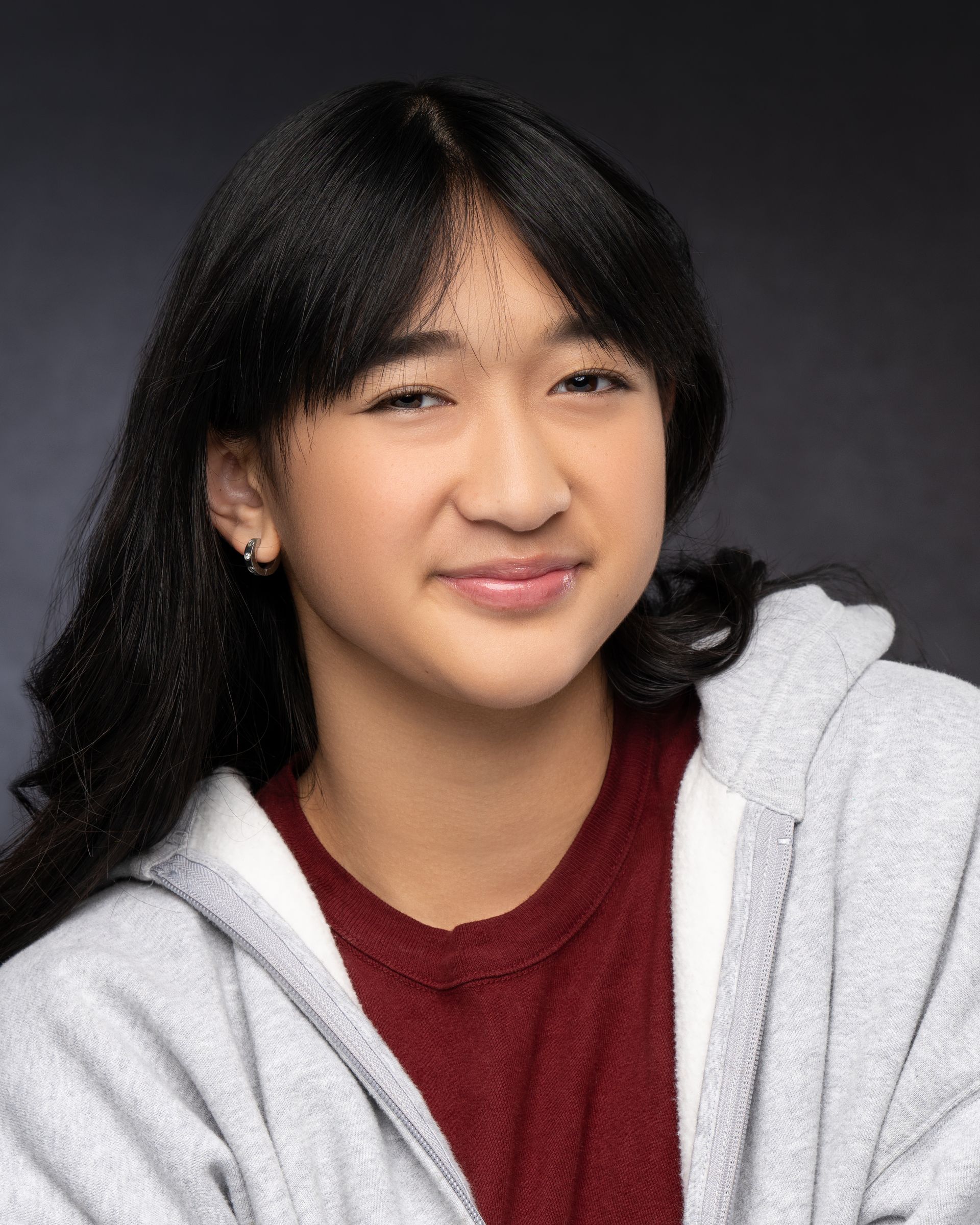 Close-up portrait of a person wearing a gray fleece hoodie over a maroon shirt, with dark wavy hair, set against a dark studio background.