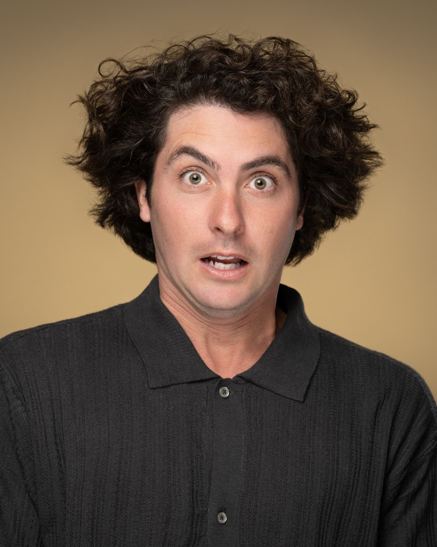 Man with curly dark hair and surprised expression, wearing dark button-up shirt against a gold background.