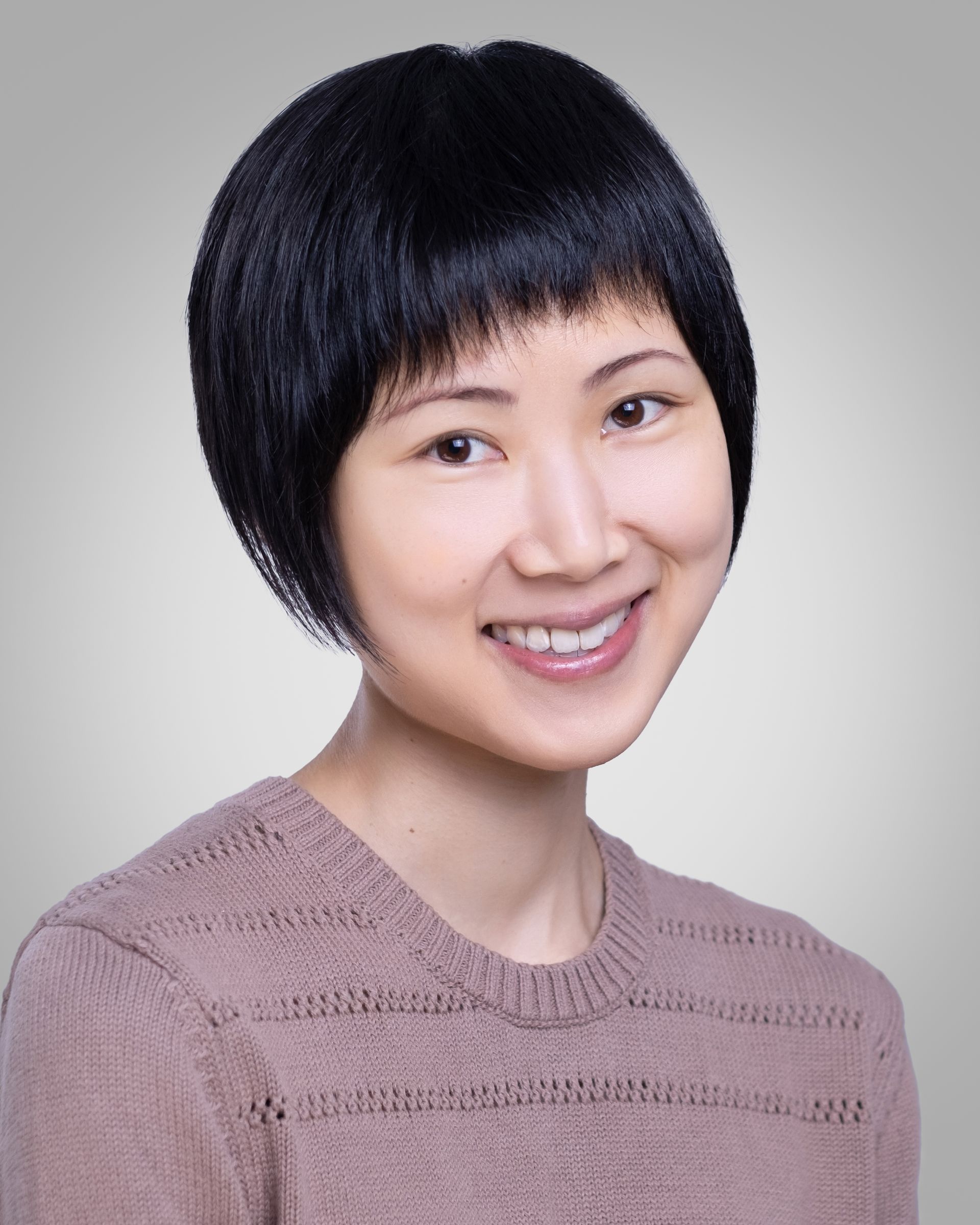 Professional Headshot of a person wearing a light mauve knit sweater with horizontal textured patterns, featuring a round neckline. The individual has short, straight black hair styled in a neat bob cut. The background is a plain light gray.