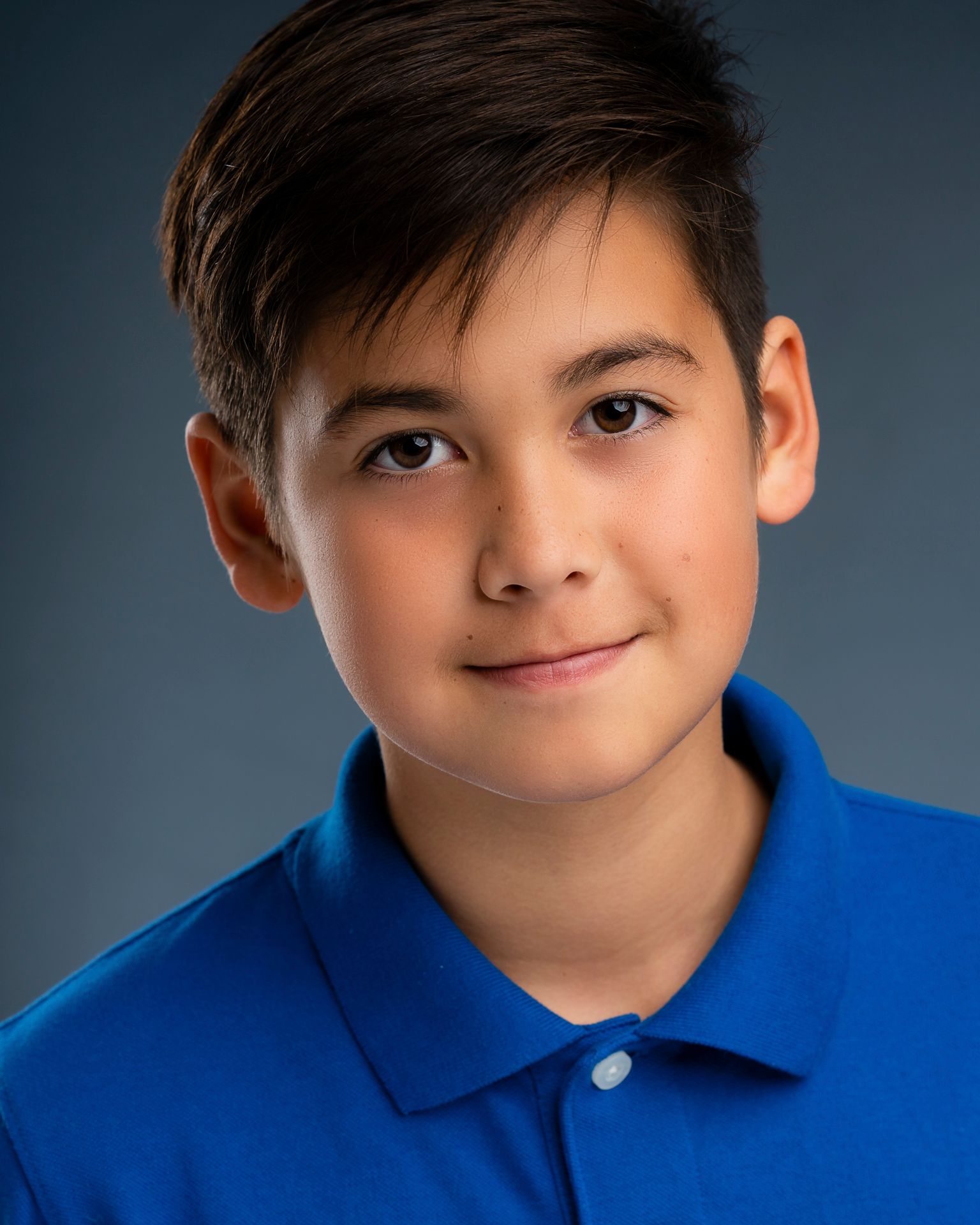 Person wearing a light blue button-up shirt with a dark inner collar detail and a chest pocket, posed against a plain dark gray background. The hair is neatly styled, and the lighting is soft and even, creating a professional studio portrait look.