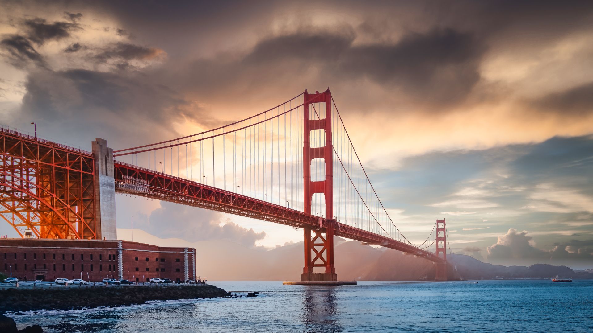 Golden Gate Bridge at sunset with dramatic clouds in the sky, viewed from the shoreline with calm blue water in the foreground.