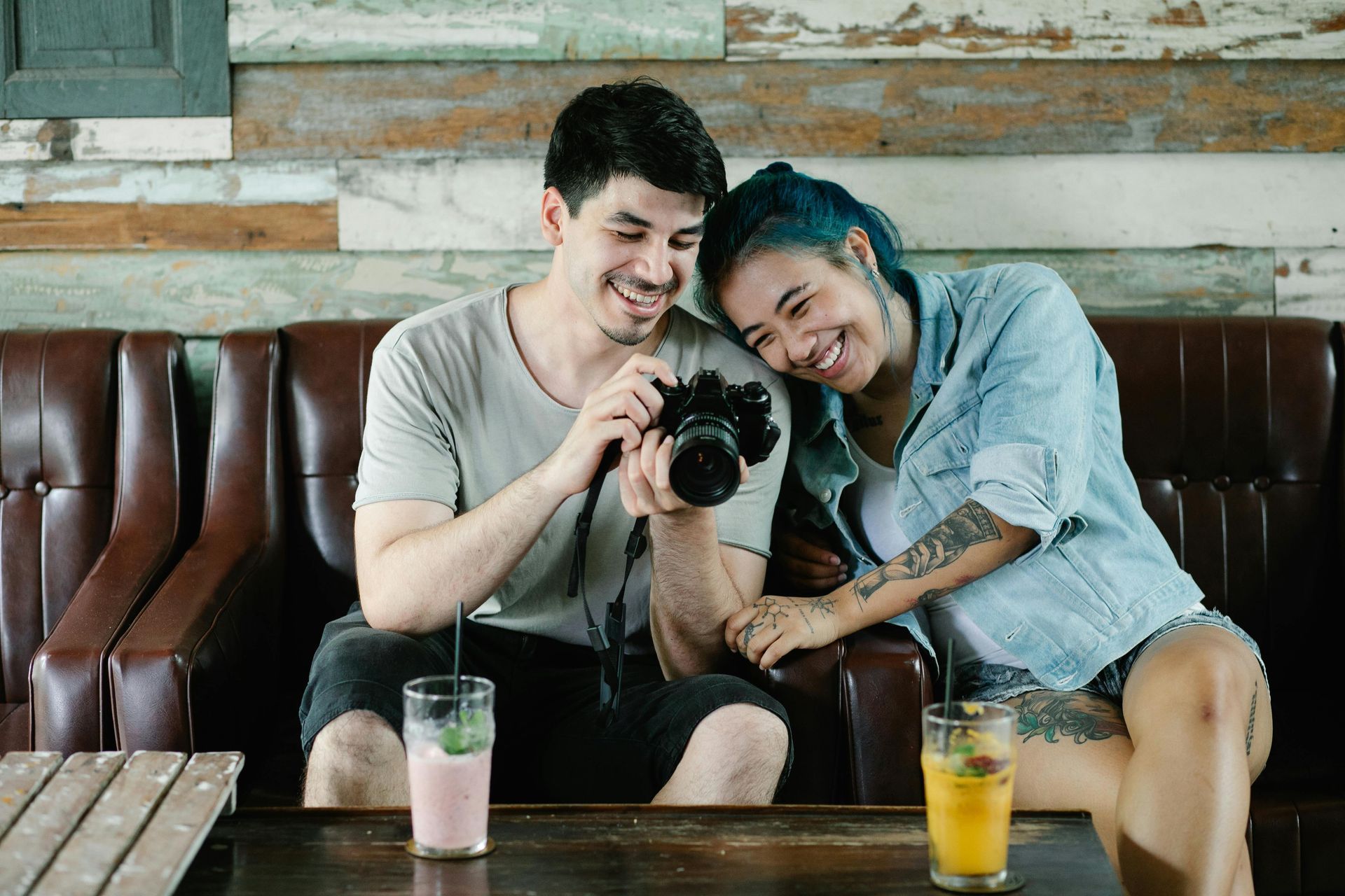 Couple smiles while looking at photos on a camera, sitting on a leather sofa. Drinks on a table in front.