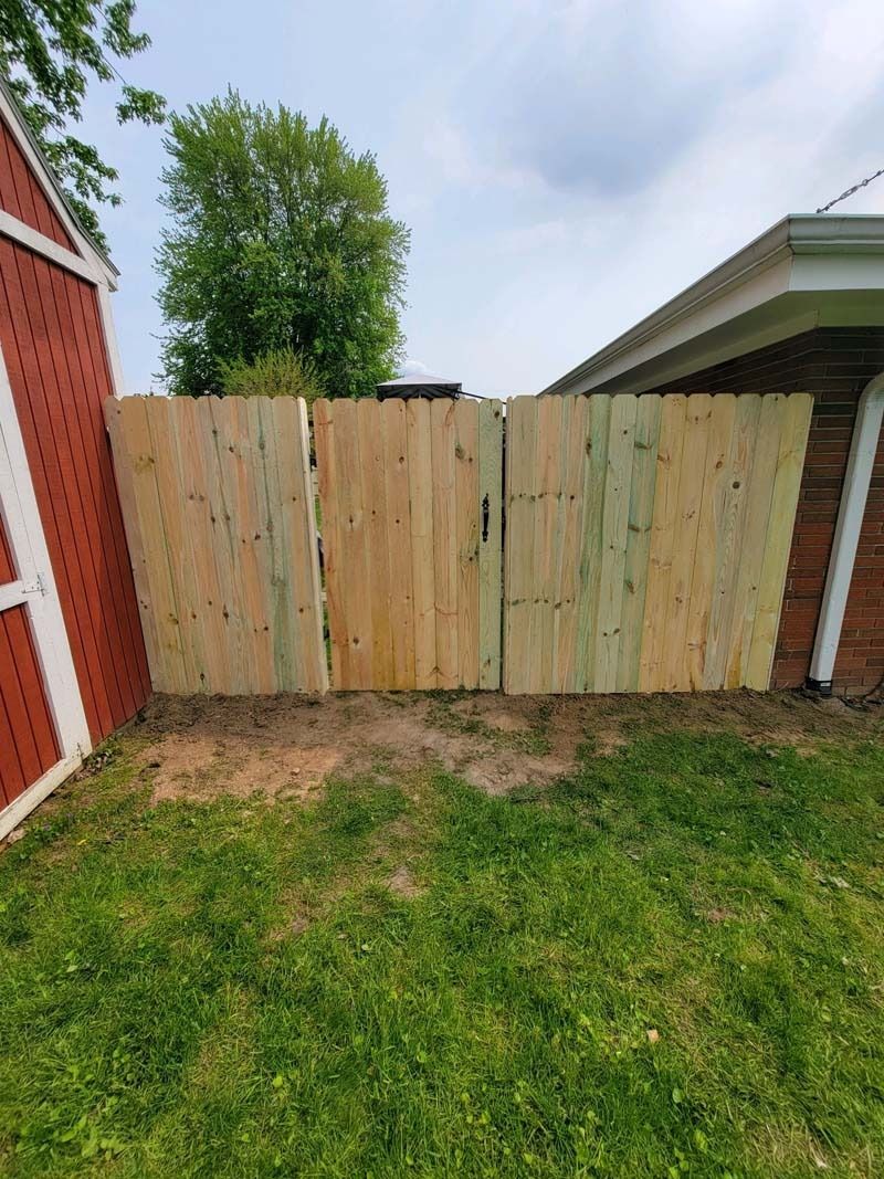 Wooden gate in a yard next to a red shed and a white-trimmed house. Green grass in the foreground.