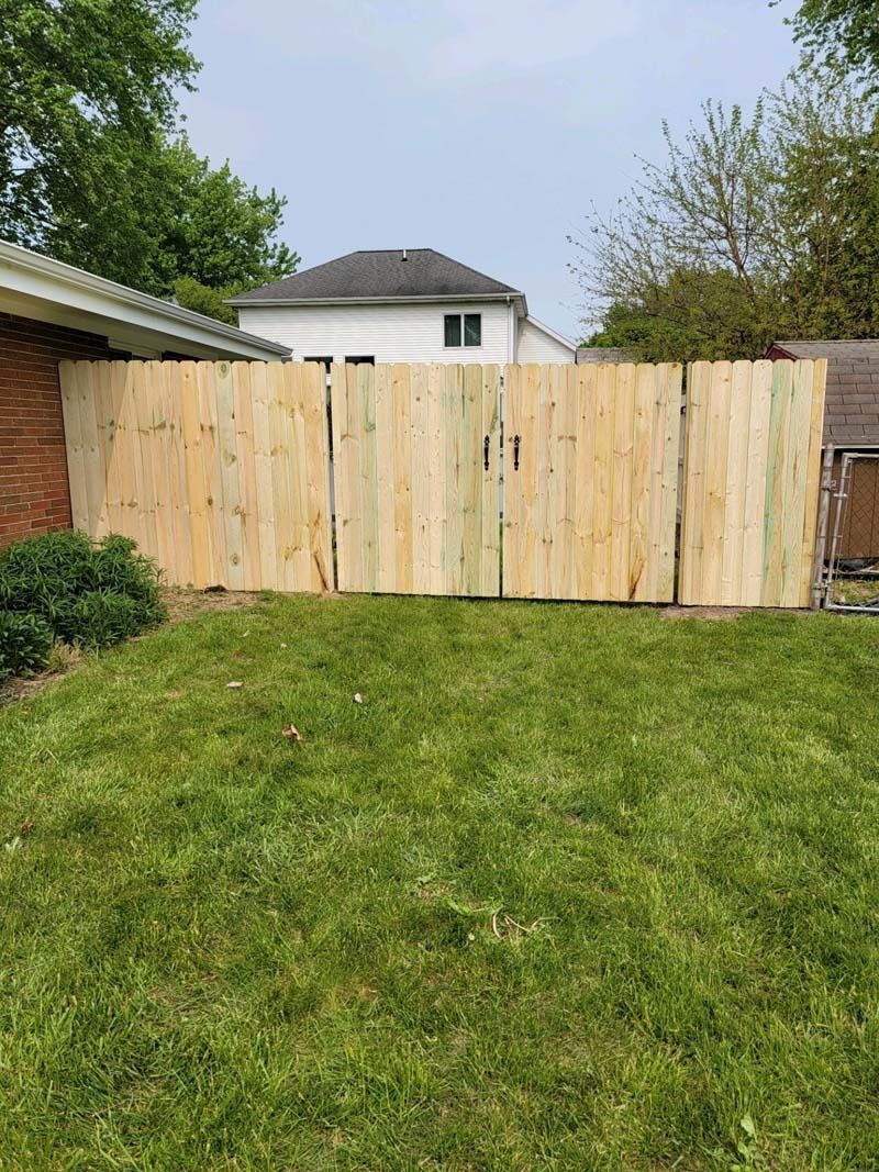 Wooden privacy fence in a backyard with green grass, trees, and a house in the background.