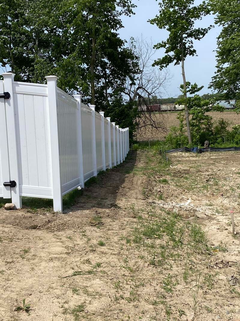 White vinyl fence along a dirt and grassy area, trees in background.