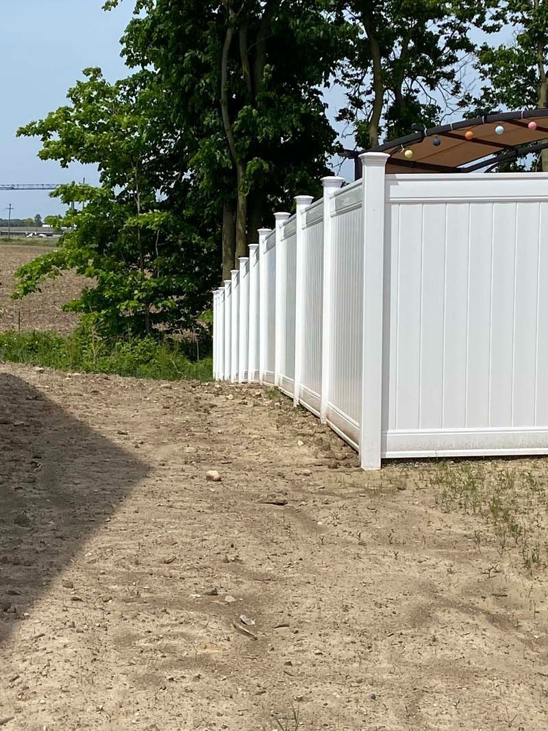 White vinyl fence alongside a dirt path, with trees and a building in the background.