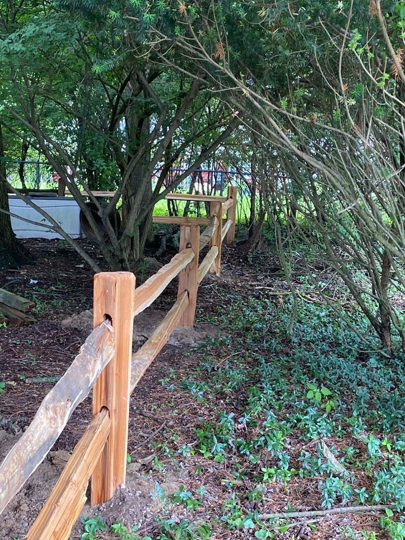 Wooden split-rail fence in a shaded, wooded area. Brown fence posts and rails. Green foliage surrounds.