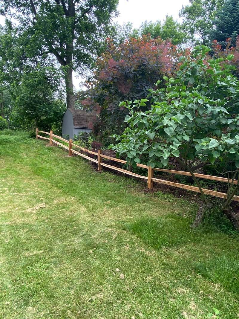 Wooden split-rail fence borders a grassy area with trees and bushes in the background.