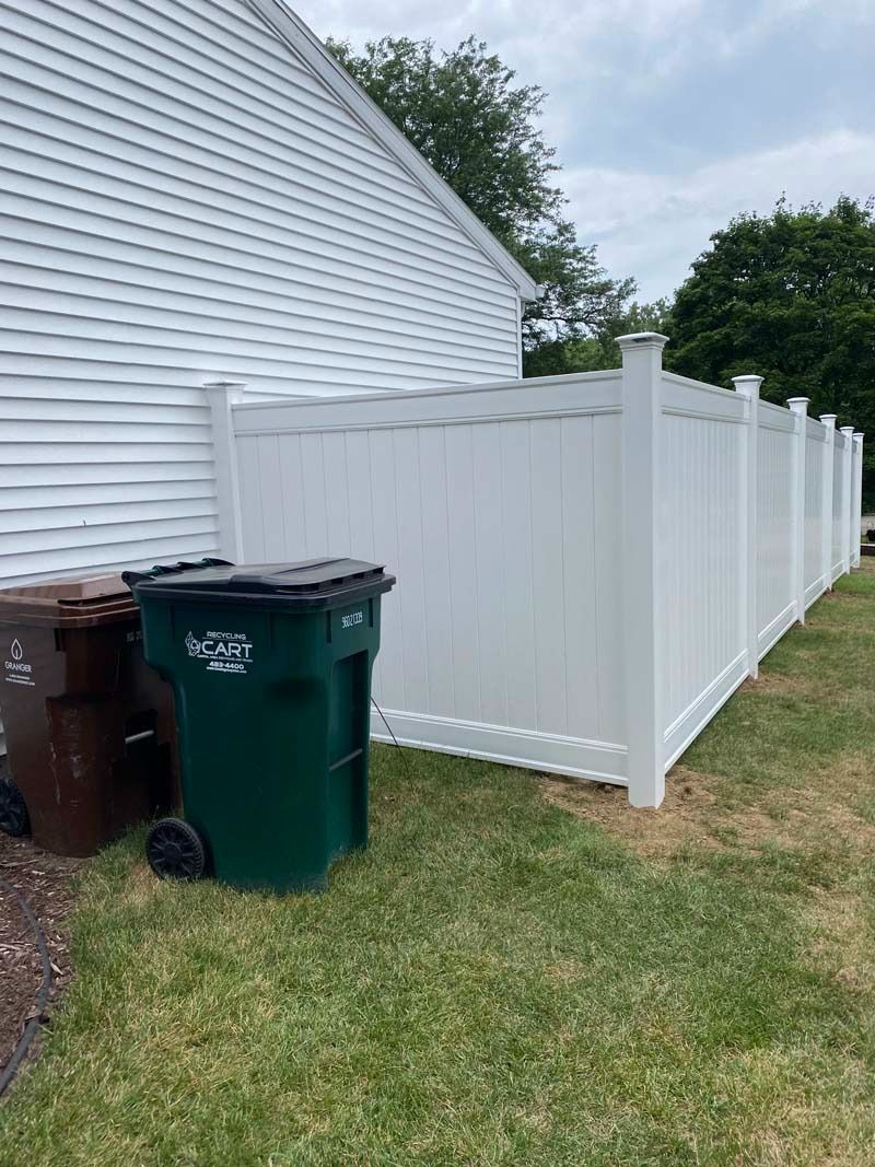 White fence enclosing trash cans next to a house with white siding. Green grass and cloudy sky.