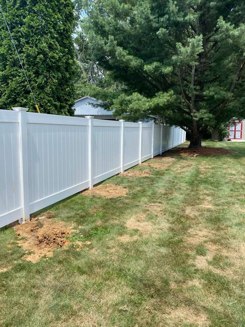 White vinyl fence along a grassy backyard, tree on the right, blue sky visible.