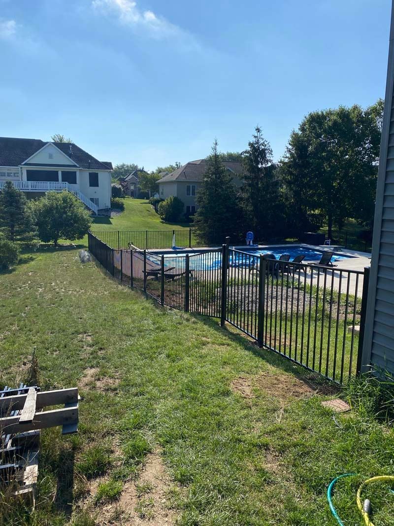 Backyard with pool, black fence, green grass, blue sky, and houses in the background.