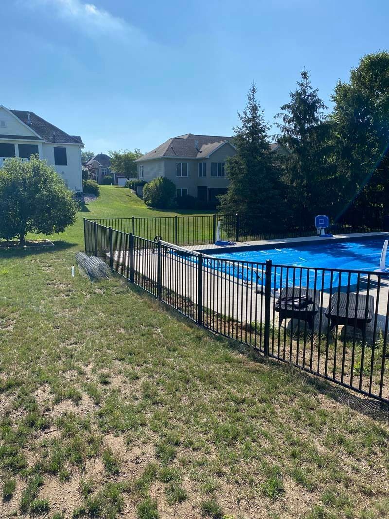 Black fence surrounds a blue-covered pool in a sunny backyard with houses and trees.