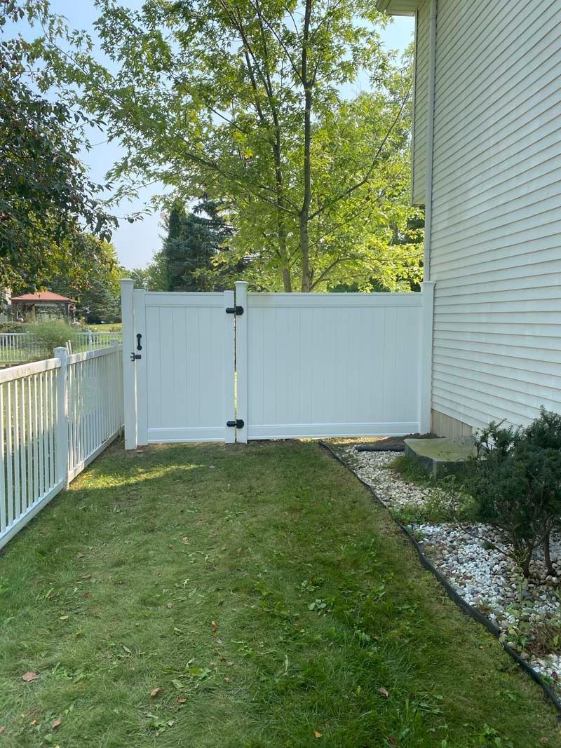 White vinyl fence with gate next to a house. Green grass in foreground and trees in the background.