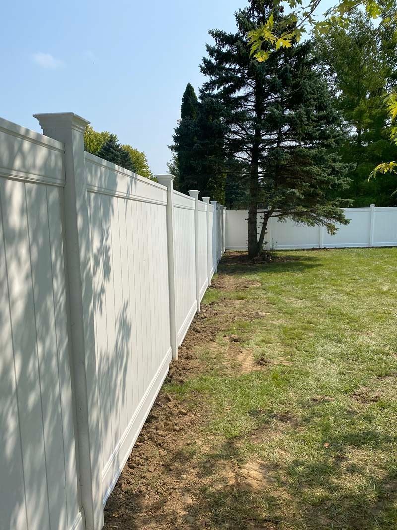 White vinyl fence in a backyard with green grass and trees under a blue sky.