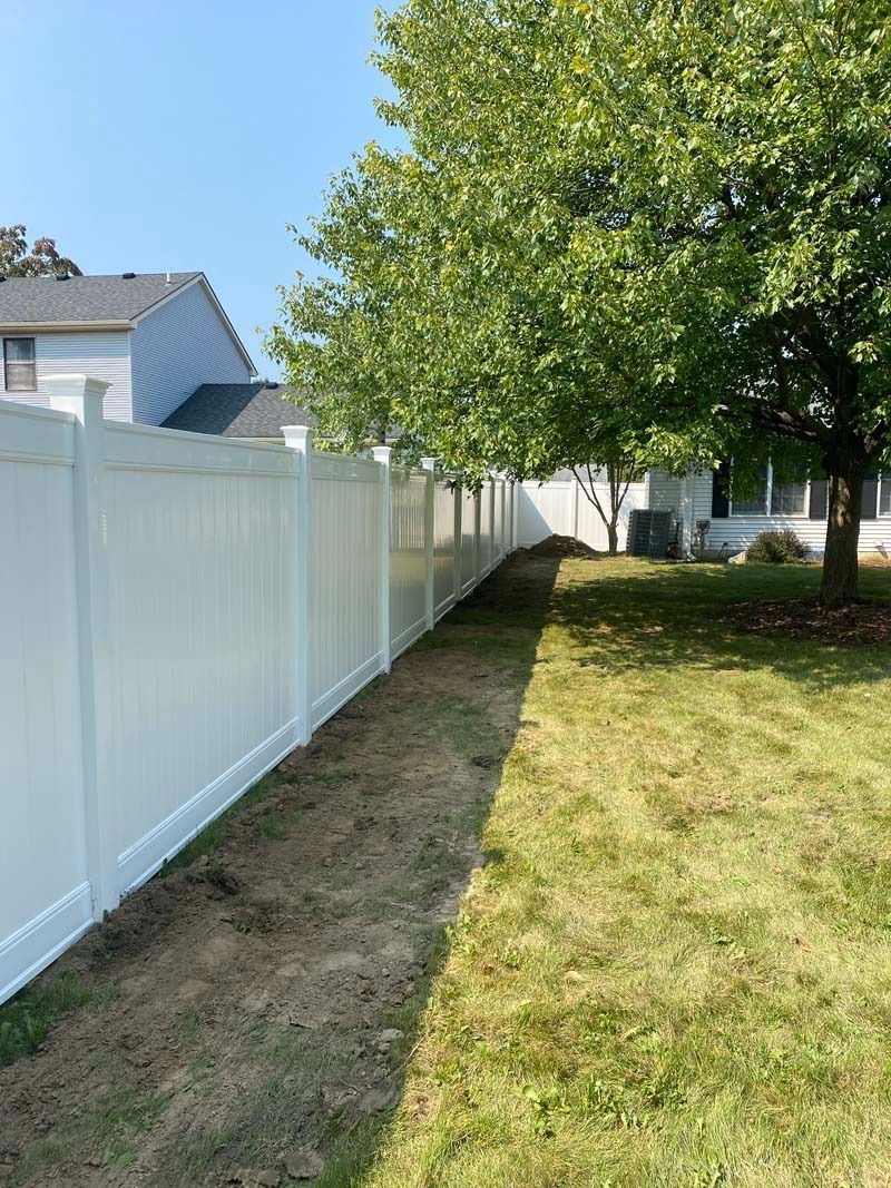 White vinyl fence in a backyard, adjacent to grass and a tree, on a sunny day.