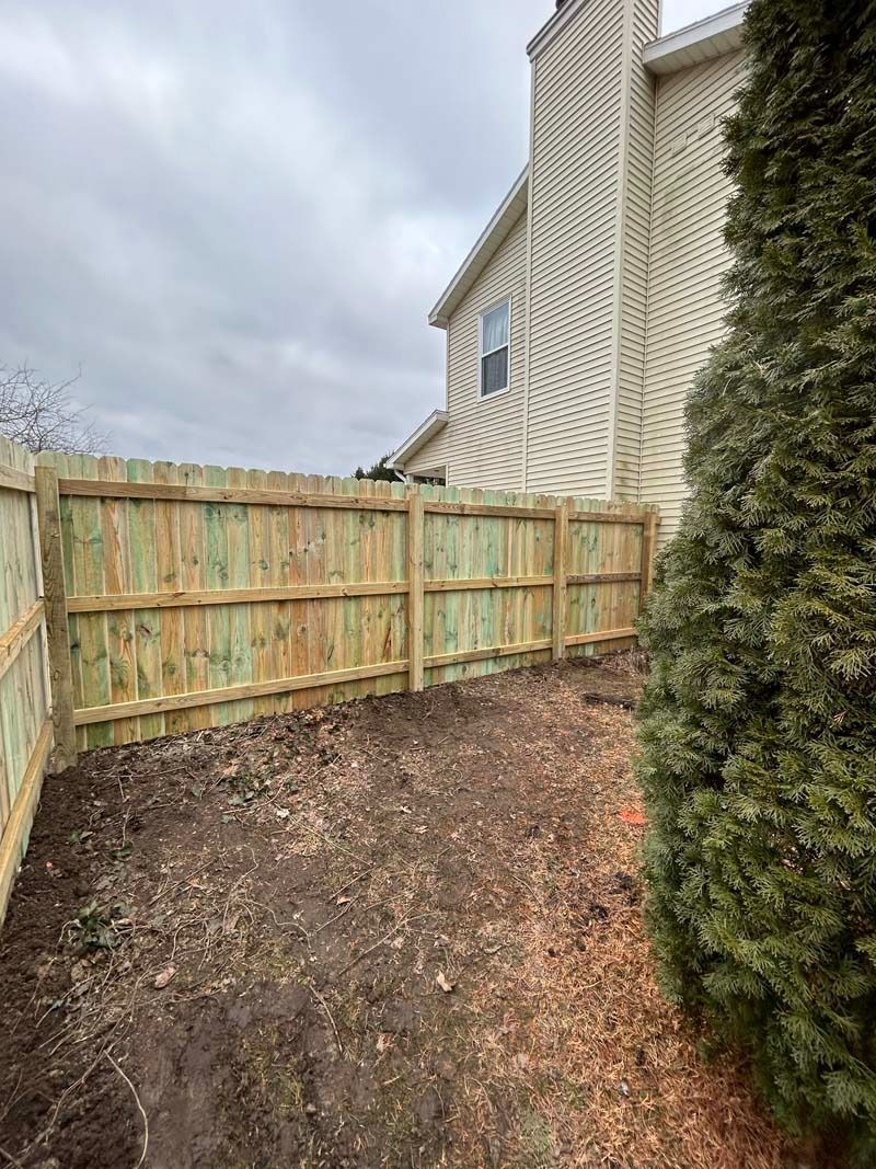 Wooden fence bordering a yard with brown soil, next to a house with a decorative facade, and a green bush.