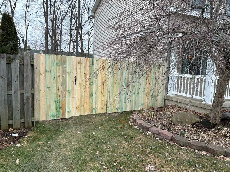 A wooden fence with new, lighter wood next to an old, gray fence by a brick building and a porch.