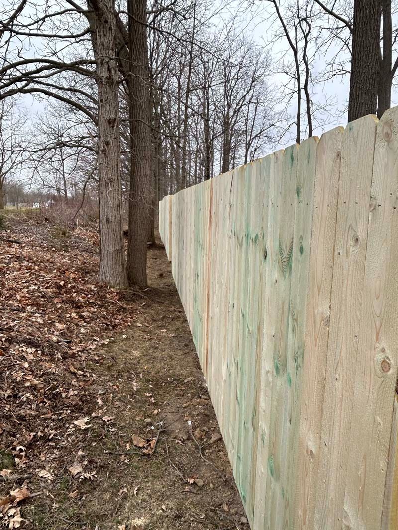 A light-colored wooden fence alongside a path in a wooded area with trees and fallen leaves.