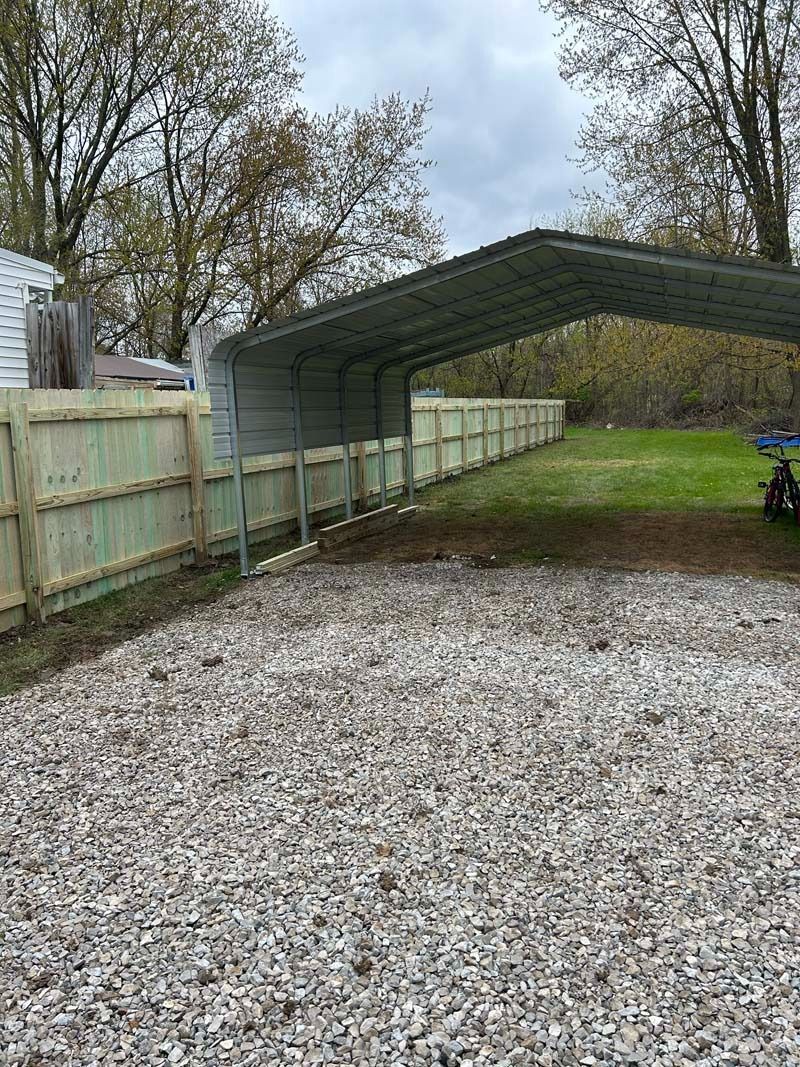 Gravel driveway next to a wooden fence and metal carport in a grassy yard.