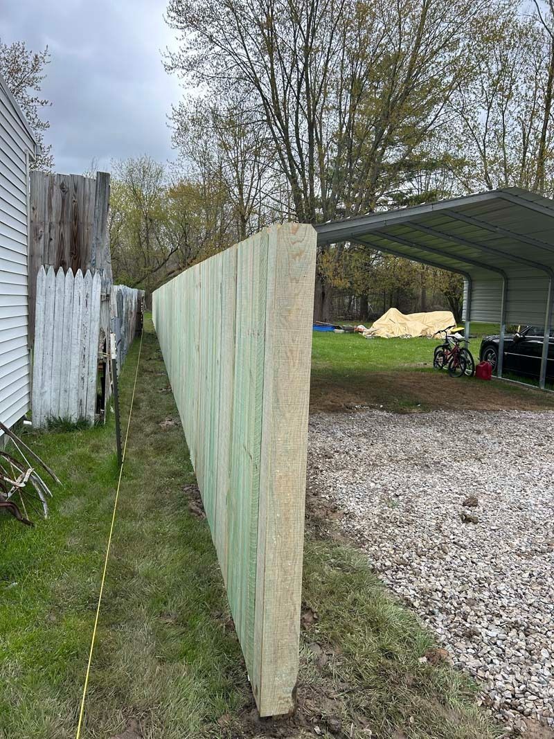 A long, green-toned wooden fence runs alongside a gravel driveway, trees in the background.