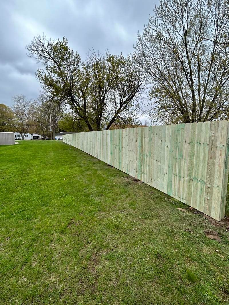 Newly built wooden fence bordering a grassy yard, trees in the background under a cloudy sky.