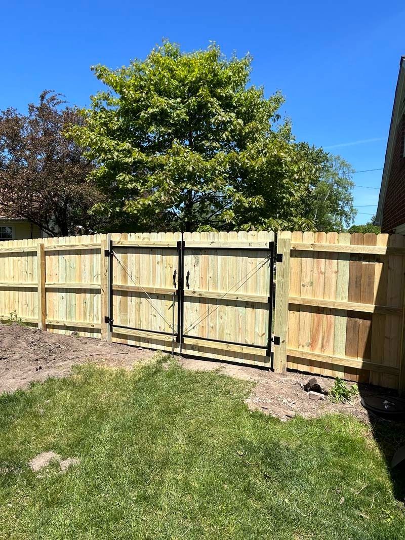 Wooden fence with black gate on a sunny day, grass in the foreground, trees in the background.
