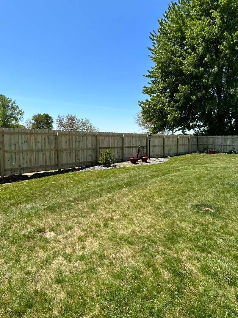 A green lawn with a wooden fence under a bright blue sky and a lush green tree.
