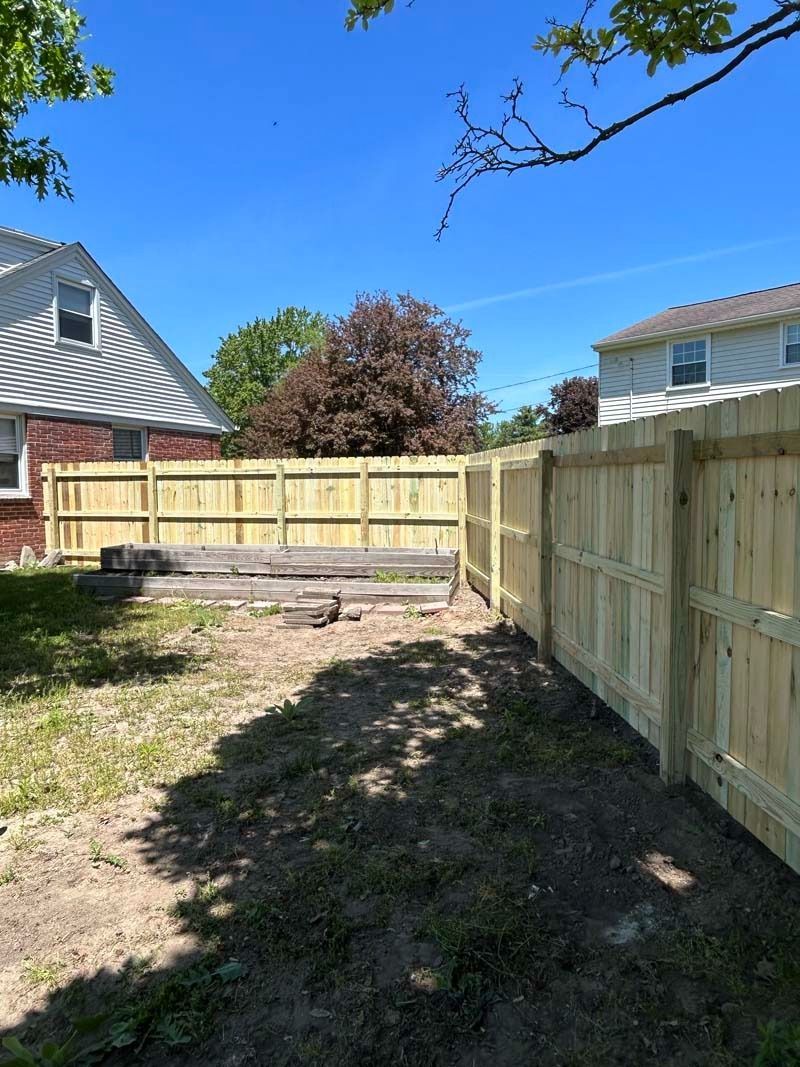 A wooden fence surrounds a backyard with a building, trees, and blue sky.