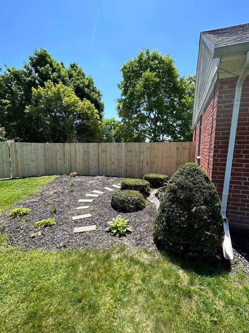 Backyard with a wooden fence, a brick house, a garden bed, and green grass.
