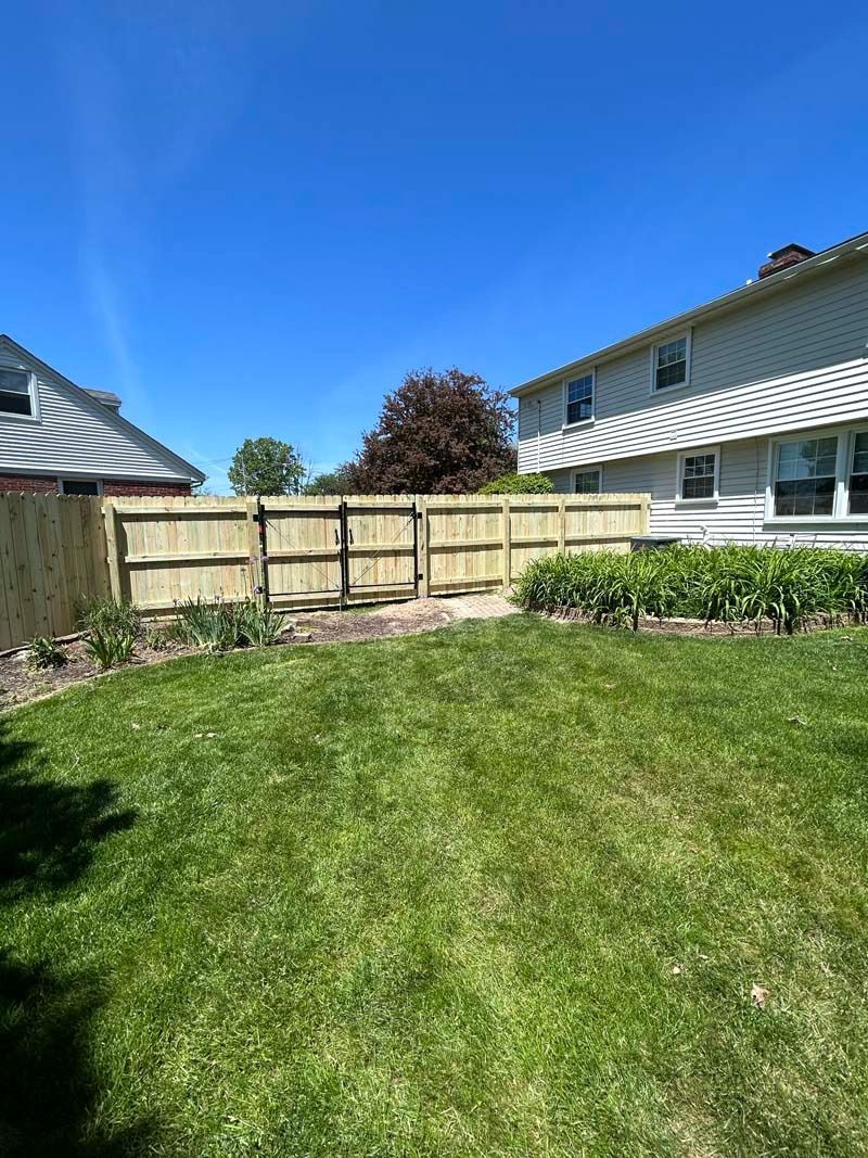 Backyard with new wooden fence under a blue sky; lawn and flowers in the foreground, house in the background.